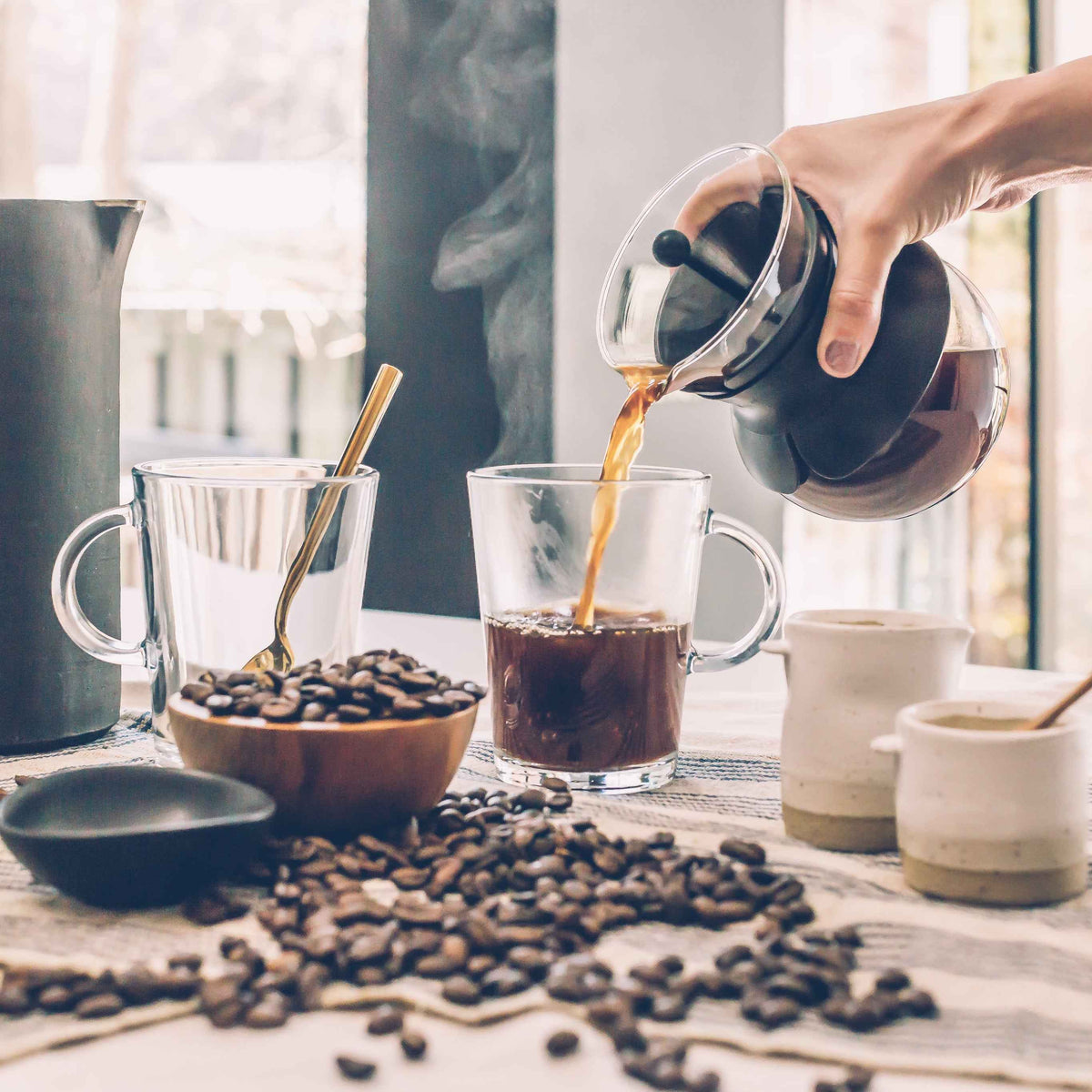 Person pouring coffee from a glass pot into a clear mug on a table with coffee beans and a coffee pot.